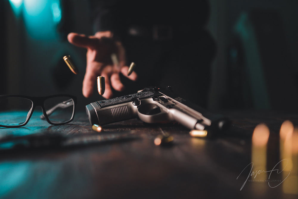 An atmospheric photograph titled 'The Shot' by Jason C. of Northern Ballistics, showcasing a handgun resting on a wooden surface with bullets suspended in mid-air. The image is cast in moody lighting that highlights the textures of the gun and wood, with a pair of glasses and a pen lying in the background, adding to the storytelling of the shot. The Northern Ballistics watermark is elegantly placed in the lower right corner, affirming the authenticity and artistry of the work.